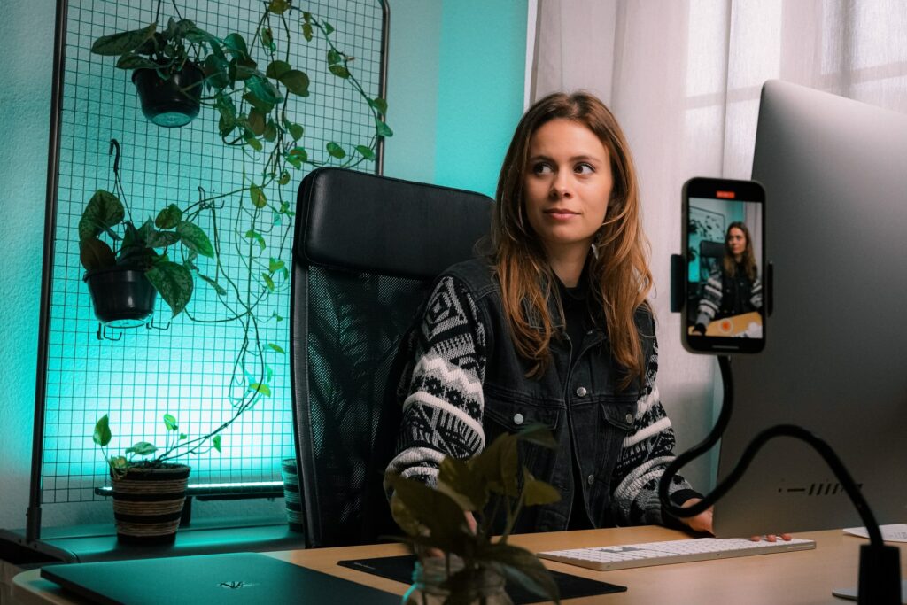 Photo of woman (light skin and long brown hair, wear a knitted, patterned sweater in grey and beige), she is sitting at a desk, in a black leather chair, in front of a computer. The background wall is turquoise.